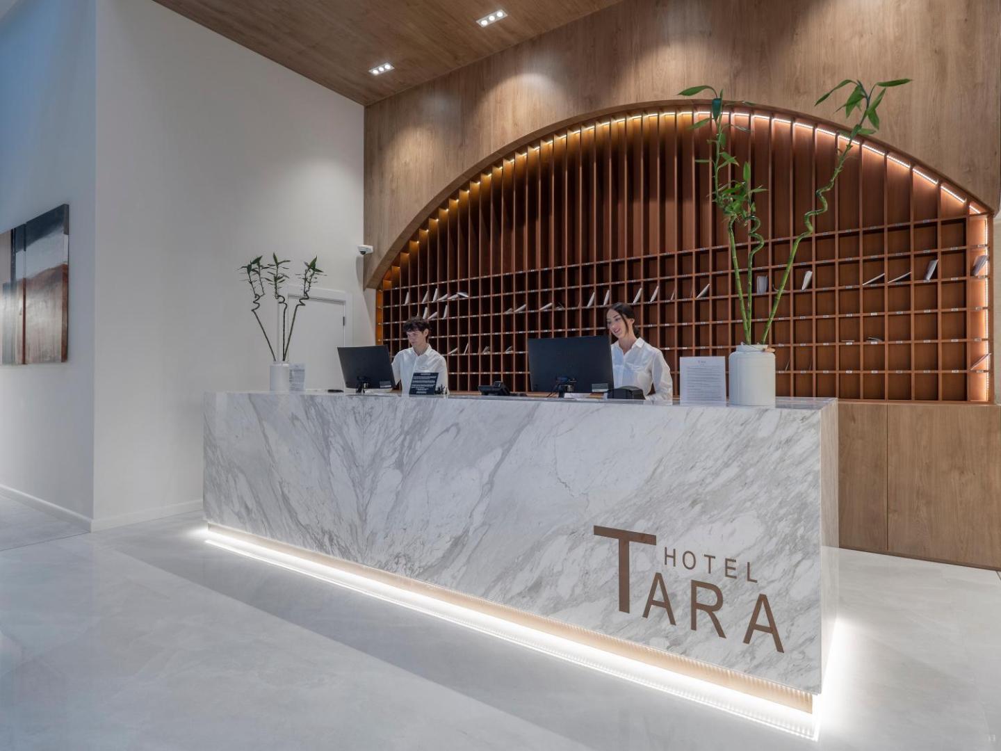 Hotel Tara recepcijas lete no marmora ar viesnīcas logo — divi darbinieki apkalpo viesus pie modernās reģistratūras