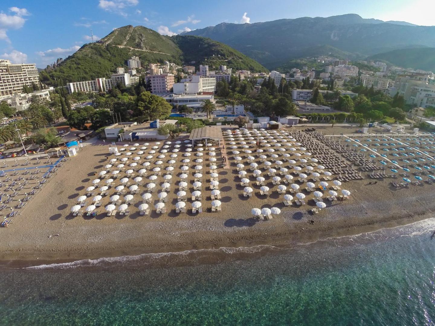 Aerofoto skats uz Hotel Tara privāto pludmali ar simtiem baltu saulessargu un kalnu fonu Bečiči kūrortā
