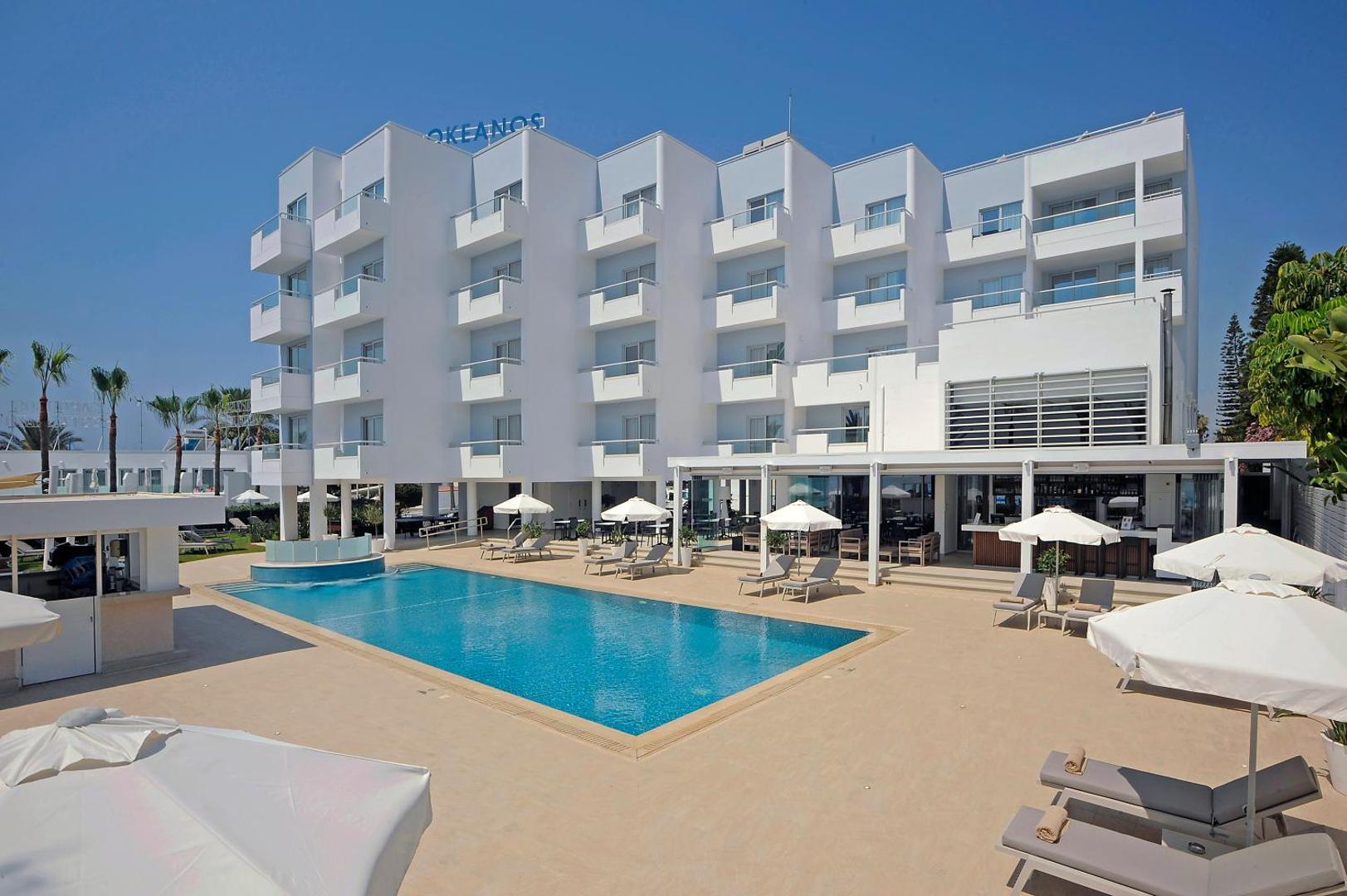 Okeanos Beach Boutique viesnīcas āra baseins Ajanapā, Kiprā — gaiša atpūtas zona ar sauļošanās krēsliem, saulessargiem un bāru pie baseina