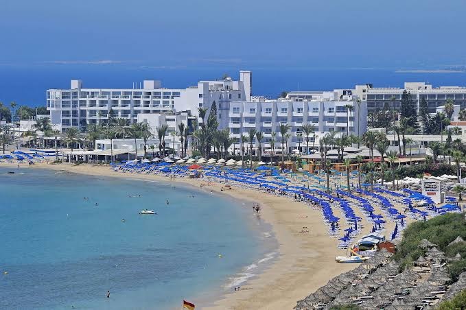 Panorāmas skats uz Okeanos Beach Boutique un Ajanapas pludmali — plaša smilšu josla ar ziliem saulessargiem un tirkīzzilu jūras ūdeni