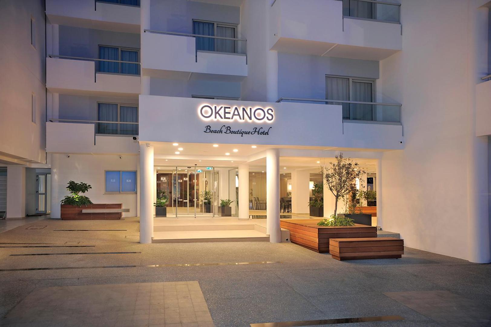 Okeanos Beach Boutique viesnīcas galvenā ieeja vakarā — moderna balta fasāde ar izgaismotu logo un koka dizaina elementiem