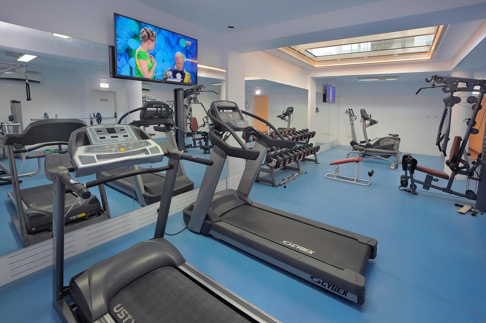 Okeanos Beach Boutique fitness centrs ar Cybex skrejceļiem un svariem — gaiša trenažieru zāle ar spoguļiem un zilu grīdas segumu