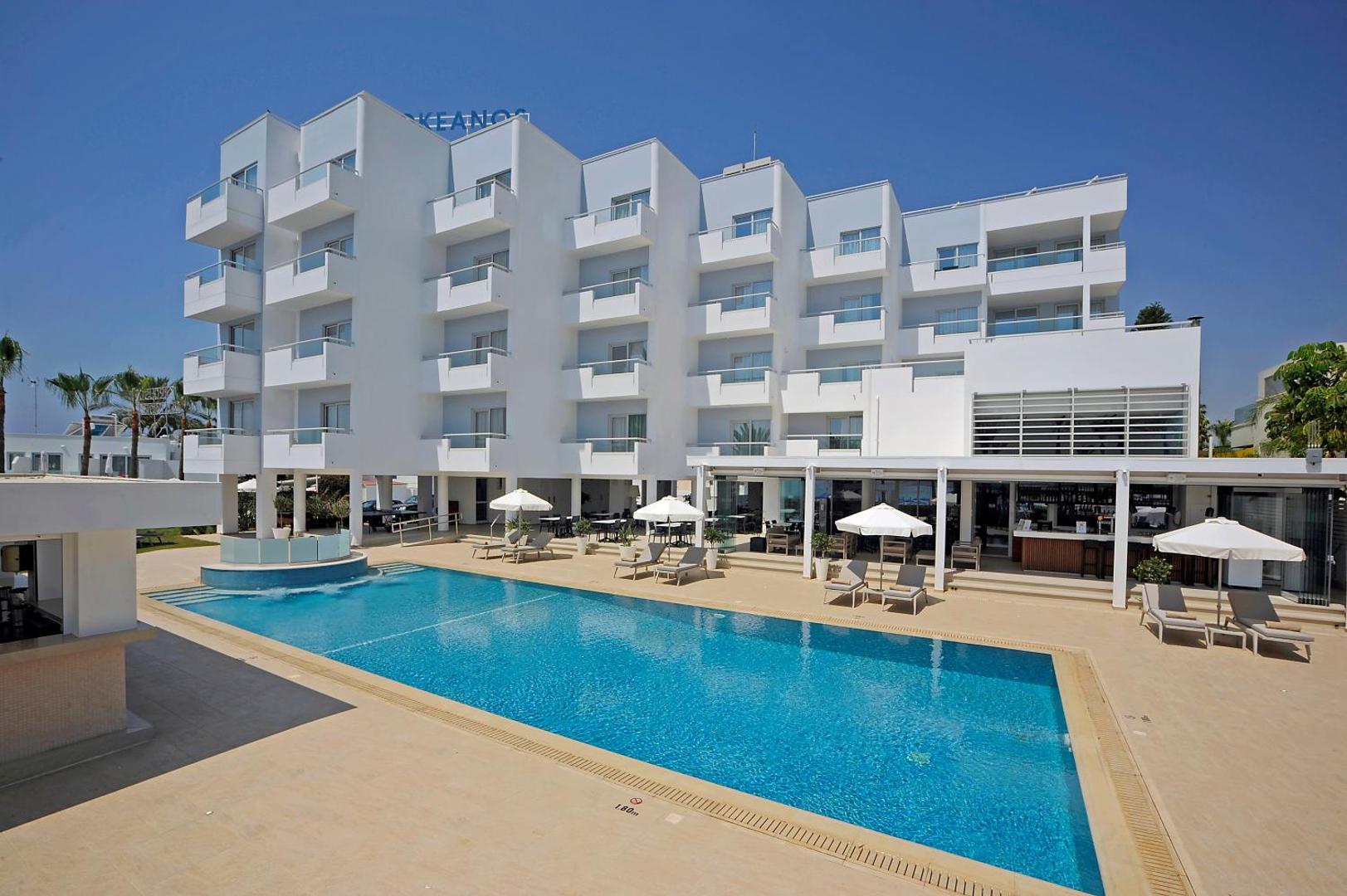 Okeanos Beach Boutique viesnīcas modernā fasāde ar balkoniem un āra baseinu Aijanapā, Kiprā — gaiša arhitektūra piekrastē