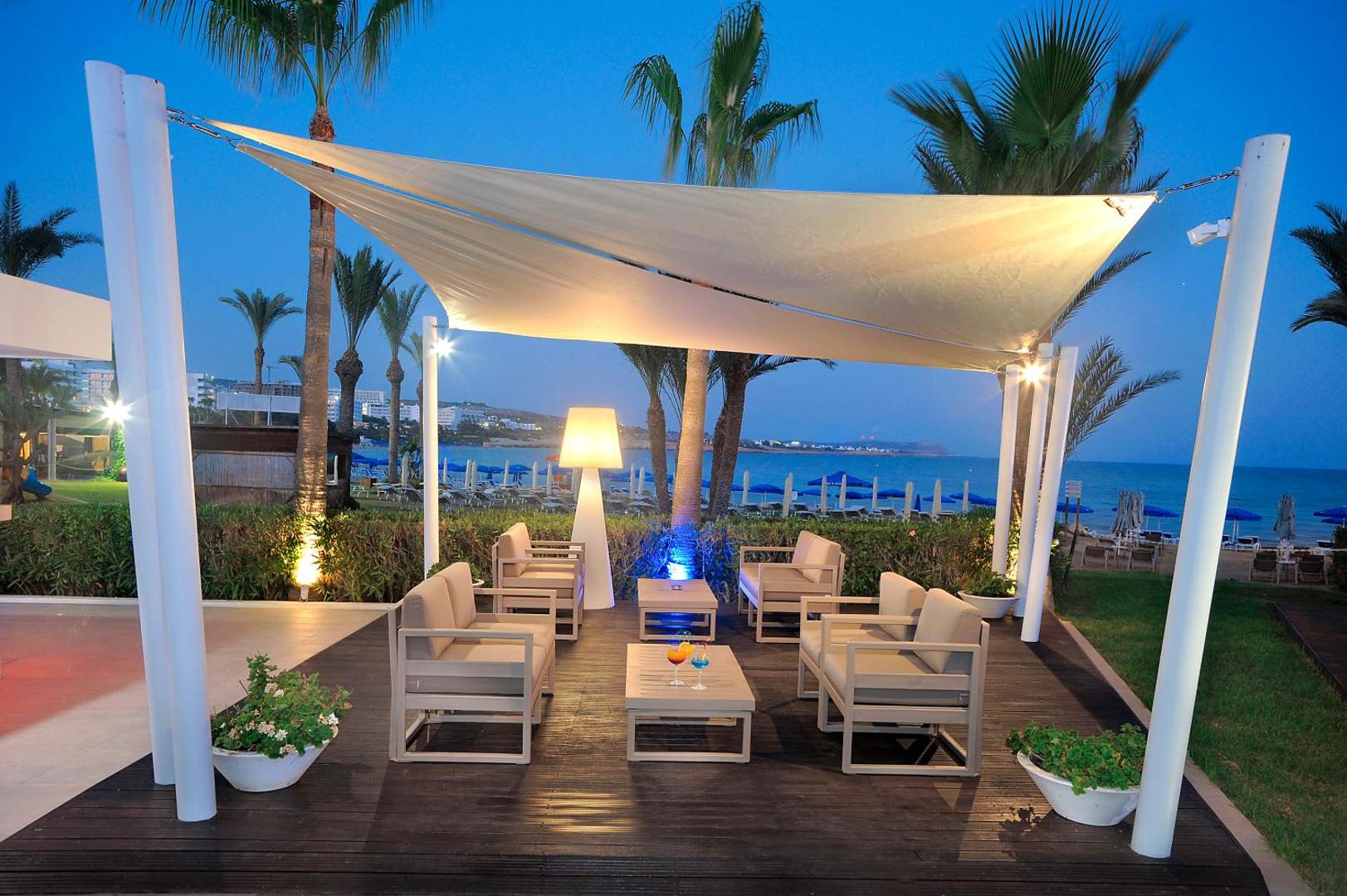 Okeanos Beach Boutique viesnīcas āra atpūtas zona ar dīvāniem zem nojumes vakarā Ajanapā, Kiprā — skats uz pludmali un palmām