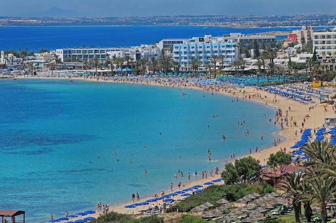 Okeanos Beach Boutique panorāmas skats uz plašo smilšu pludmali Ajanapā — tirkīza krāsas jūra, saulessargi un viesnīcas ēkas piekrastē