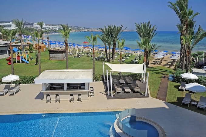 Skats no augšas uz Okeanos Beach Boutique teritoriju — baseins, bērnu rotaļu laukums un privātā pludmale ar zilajiem saulessargiem