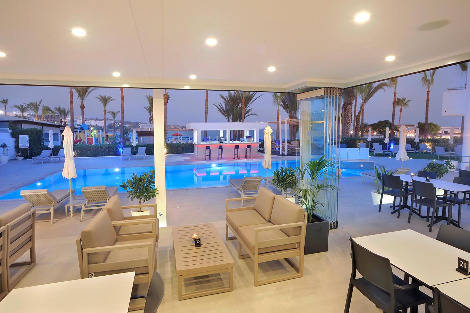 Okeanos Beach Boutique viesnīcas bārs pie baseina vakarā — moderna atpūtas zona ar dīvāniem, apgaismots baseins un palmas fonā