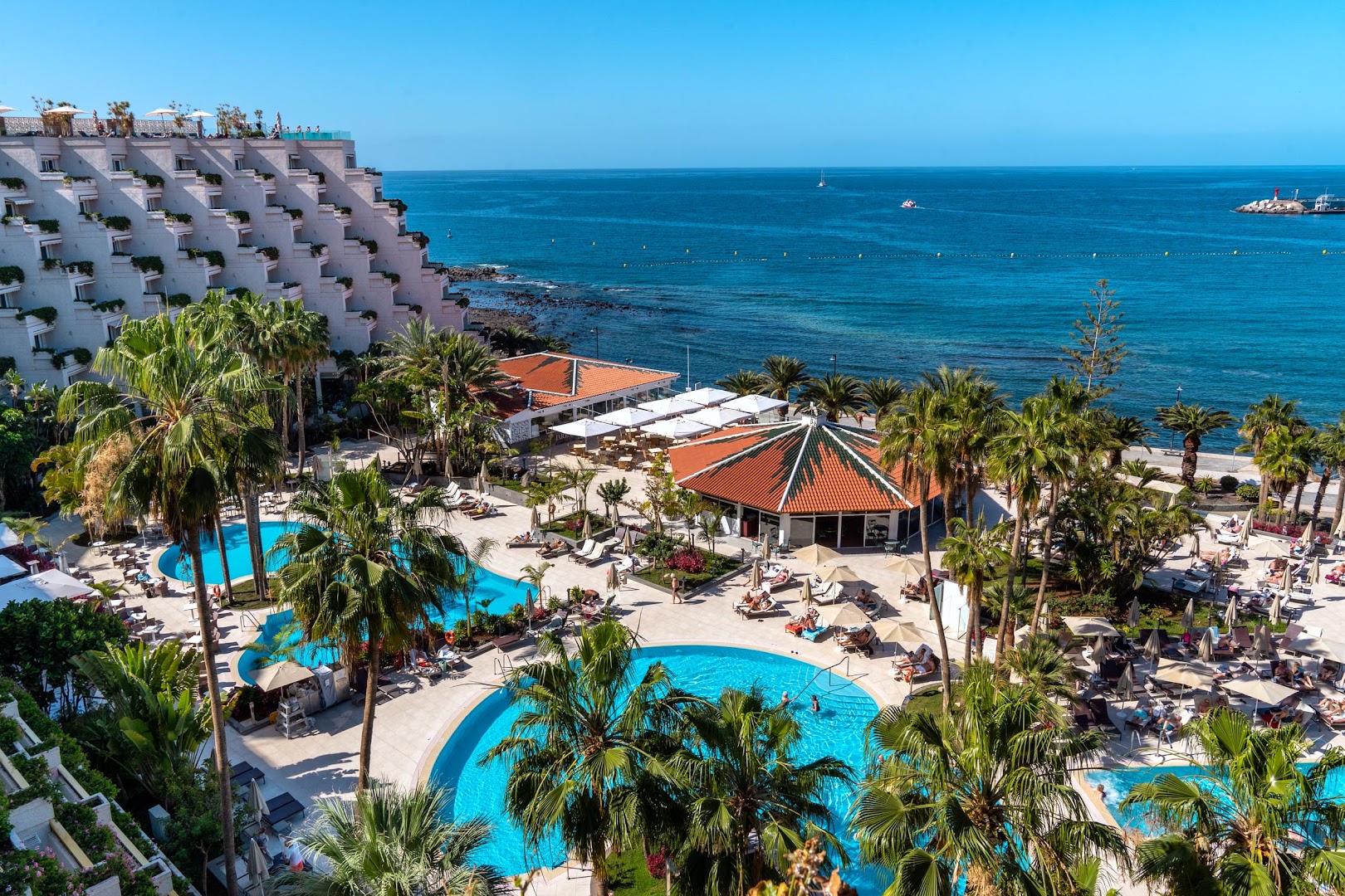 Arona Gran Hotel & Spa āra baseini ar palmām un sauļošanās krēsliem pie okeāna Tenerifē — skats uz zilo ūdeni un horizontu