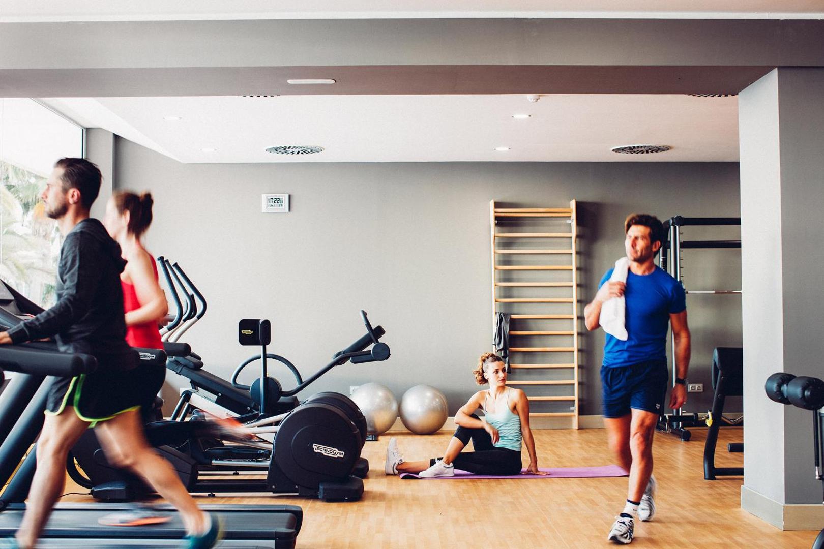 Viesi trenējas Arona Gran Hotel & Spa sporta zālē — plaša telpa ar skrejceliņiem, paklājiņiem un vingrošanas sienu