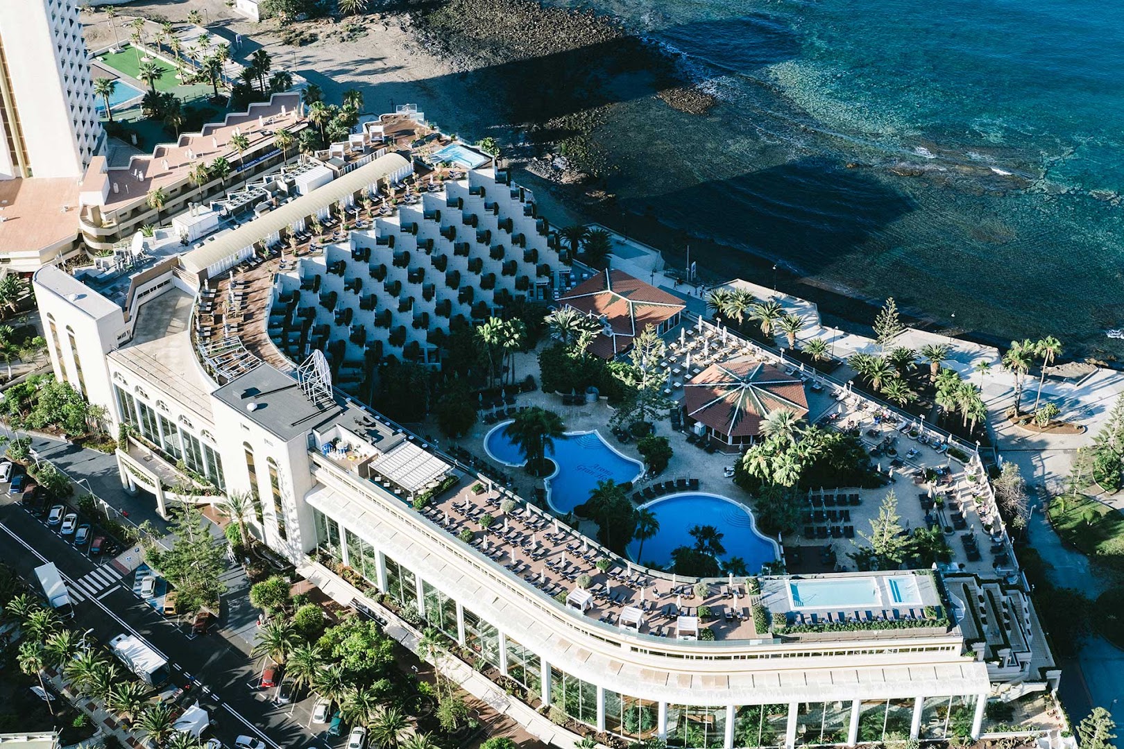 Arona Gran Hotel & Spa skats no putna lidojuma uz baseinu zonu un piekrasti Los Kristianosā, Tenerifē — redzama viesnīcas arhitektūra