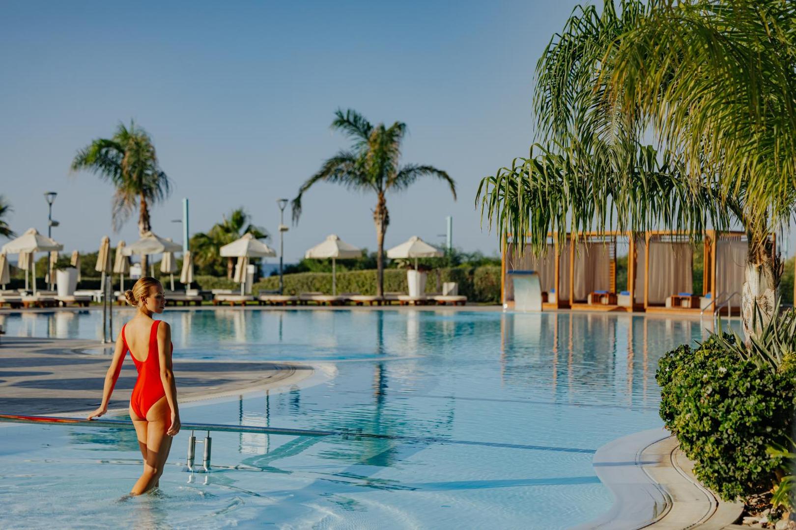 Sieviete sarkanā peldkostīmā ieiet Nissiblu Beach Resort baseinā — fonā palmas, saulessargi un atpūtas nojumes zem skaidrām debesīm