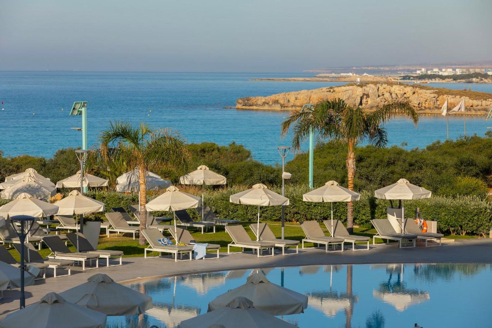 Skats no Nissiblu Beach Resort terases uz Vidusjūru un klinšaino piekrasti — priekšplānā atpūtas krēsli, saulessargi un palmas
