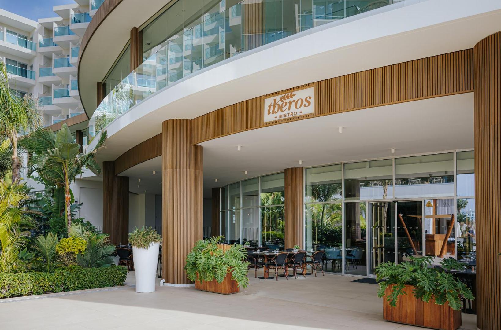 Nissiblu Beach Resort Theros Bistro ieeja ar koka apdari un stikla fasādi — moderna arhitektūra un tropu augi