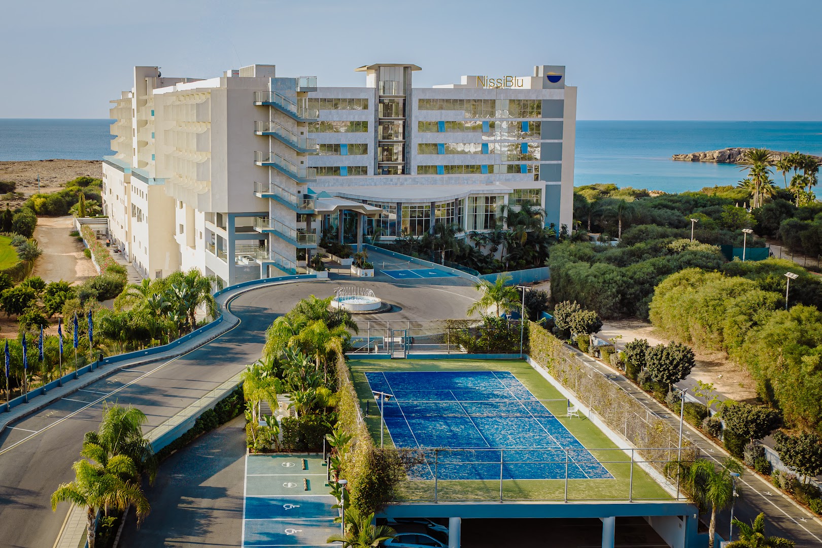 Nissiblu Beach Resort fasāde un tenisa korts Aijanapā, Kiprā — skats uz viesnīcas ieeju un zilo tenisa laukumu palmu ielenkumā