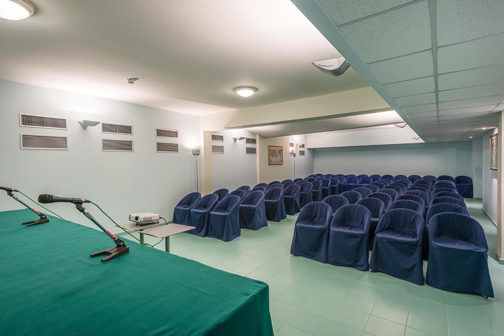 Rada Siri Hotel konferenču zāle ar zilām krēslu rindām, projektoru un mikrofonu uz zaļa galda Kalabrijas reģionā