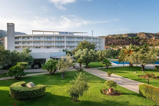 Rada Siri Hotel ēkas fasāde un plašais dārzs ar koptu zālienu un olīvkokiem Kalabrijā, Itālijā — skats uz viesnīcas teritoriju dienā