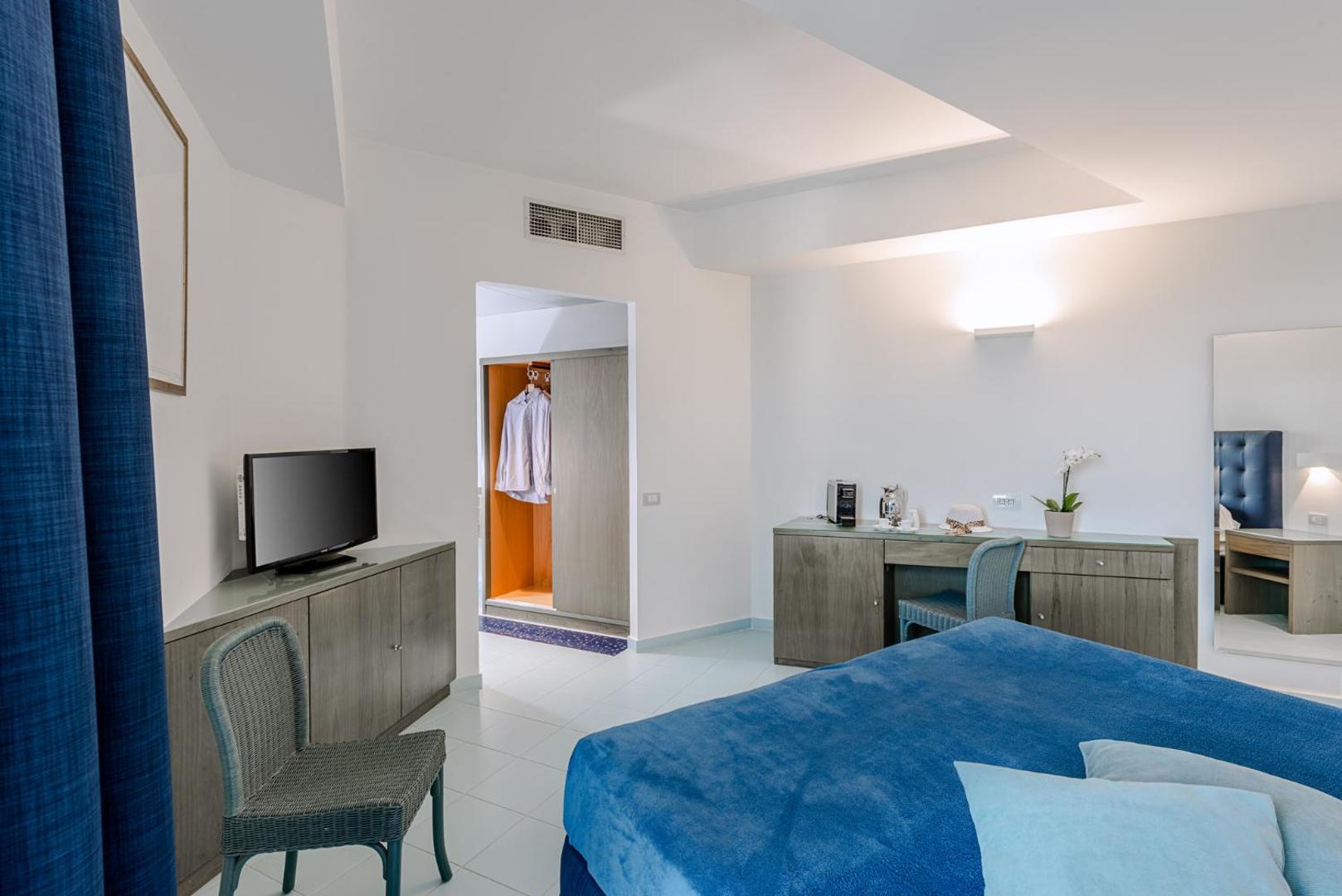 Rada Siri Hotel numura interjers ar zilu gultas pārklāju, rakstāmgaldu, televizoru un ieeju garderobē