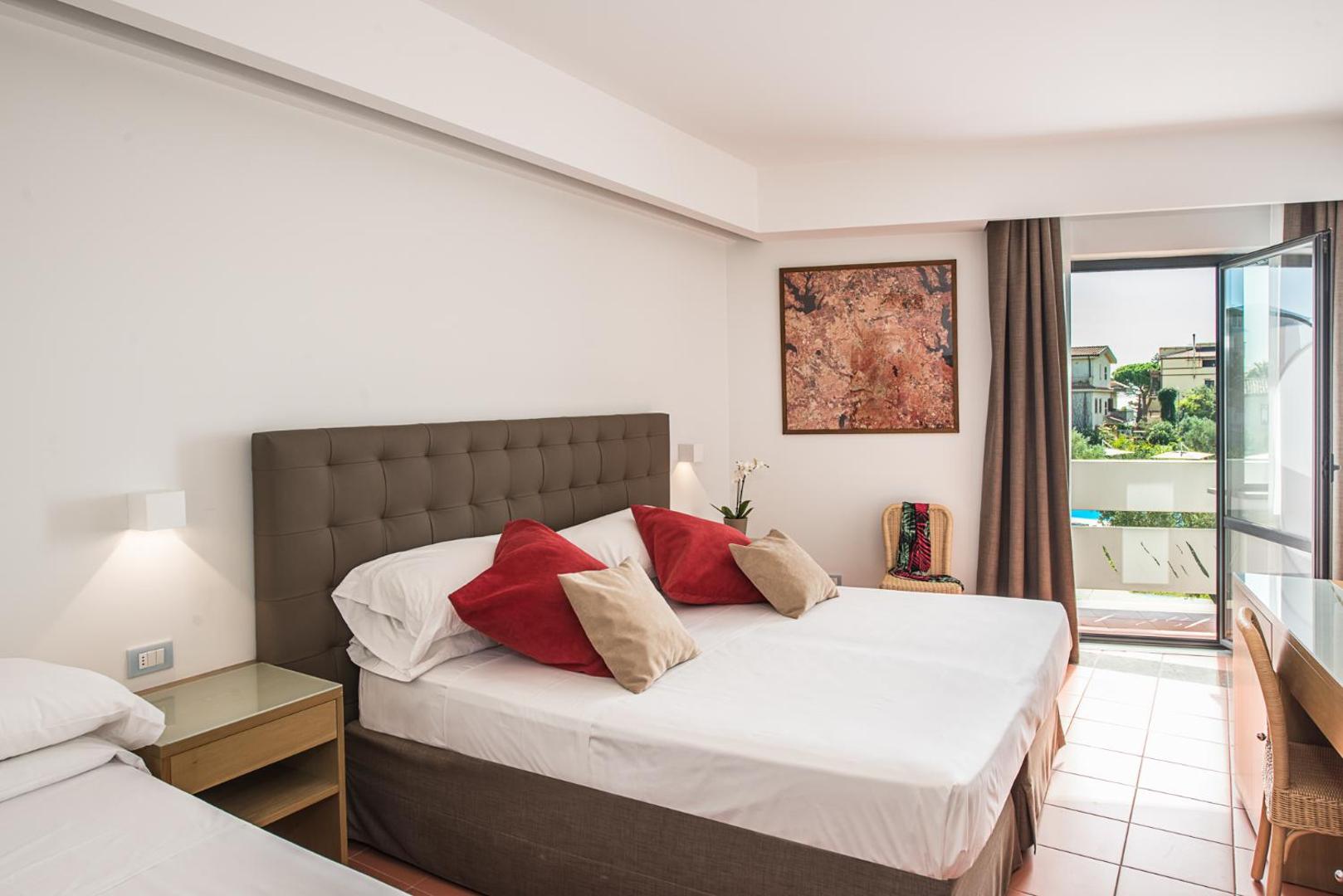 Rada Siri Hotel plašs numurs ar lielu gultu, sarkanu dekoratīvo spilvenu akcentiem un balkona durvīm