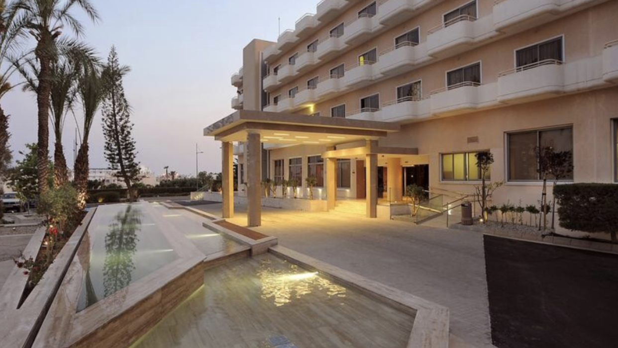 Nestor Hotel ieeja un piebraucamais ceļš vakarā — viesnīcas ēka ar balkoniem un dekoratīvu ūdens elementu priekšplānā