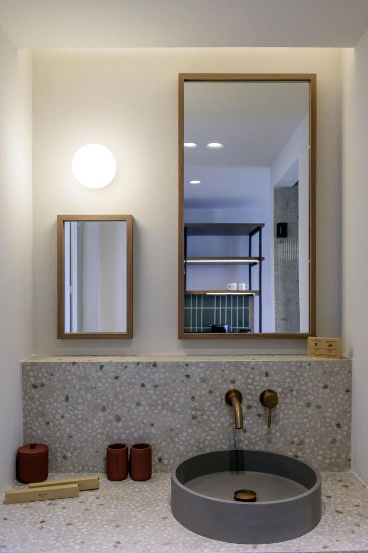 INNSiDE by Meliá Elounda vannasistabas dizains ar pelēku izlietni, terrazzo virsmu, bronzas jaucējkrāniem un diviem spoguļiem koka rāmjos.