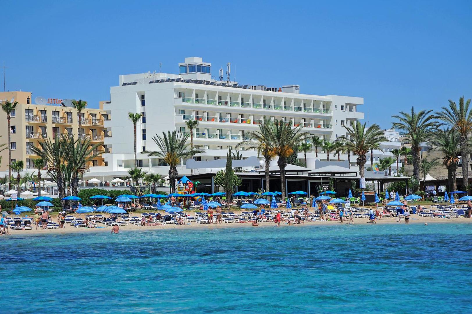 Viesnīca Nelia Beach no jūras puses — balta daudzstāvu ēka pie pašas pludmales ar palmām un saulessargiem priekšplānā