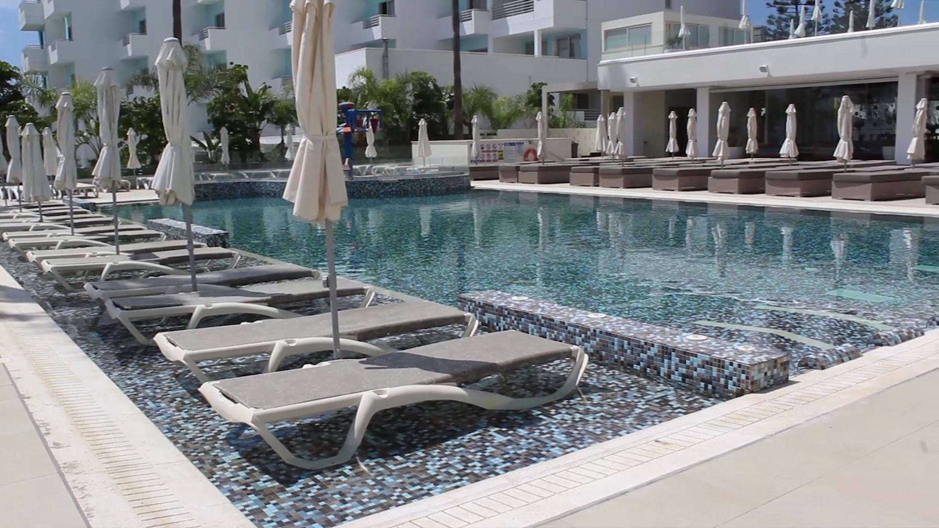 Nelia Beach Hotel sauļošanās krēsli, kas novietoti tieši seklajā baseina daļā Aijanapā — moderna atpūtas zona pie viesnīcas korpusa
