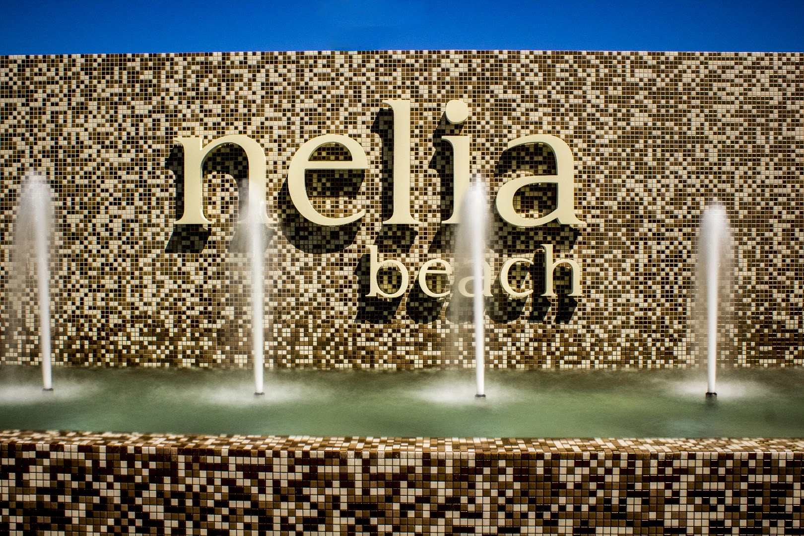 Nelia Beach viesnīcas nosaukums uz dekoratīvas mozaīkas sienas ar nelielām strūklakām priekšplānā