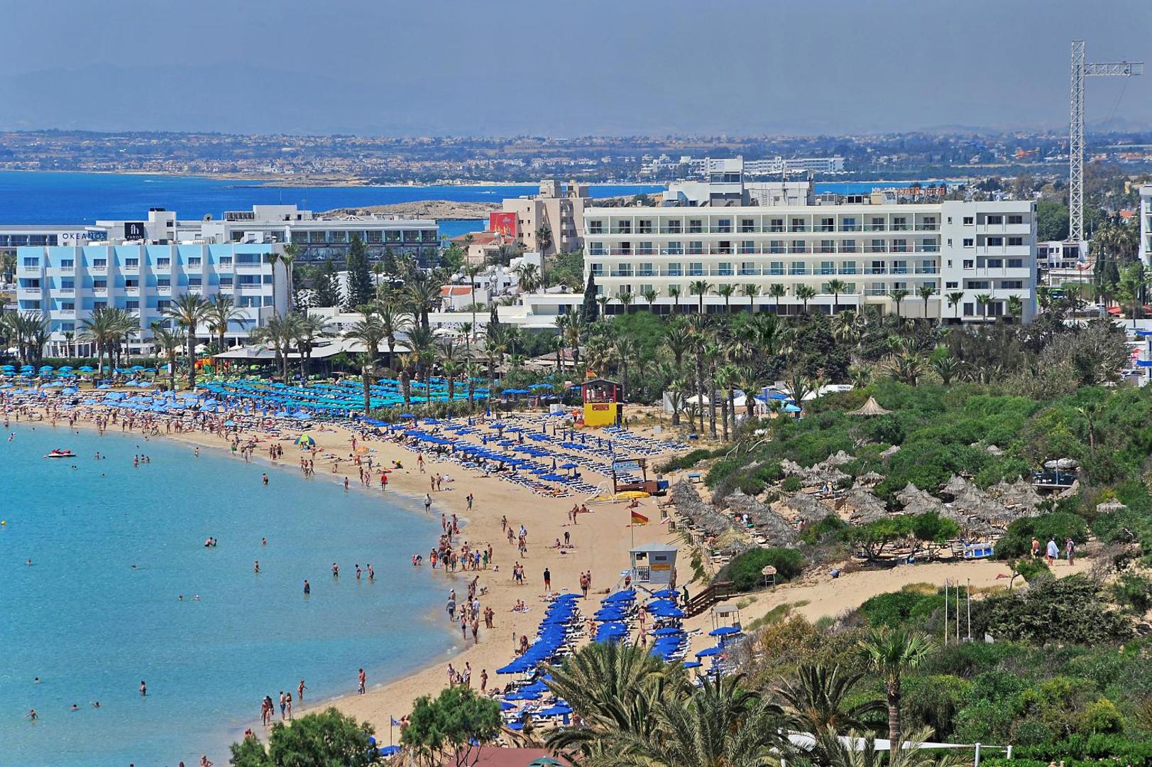 Nelia Beach viesnīcas panorāmas skats uz Ajanapas piekrasti Kiprā — plaša smilšu pludmale ar ziliem saulessargiem un pilsētas apbūvi fonā