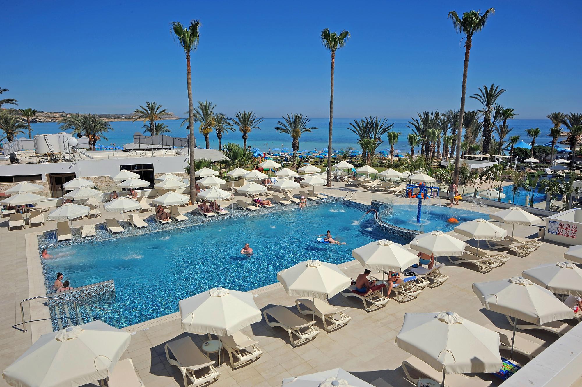 Skats no augšas uz Nelia Beach Hotel baseina zonu un atpūtas krēsliem Aijanapā — fonā redzama jūra un palmas
