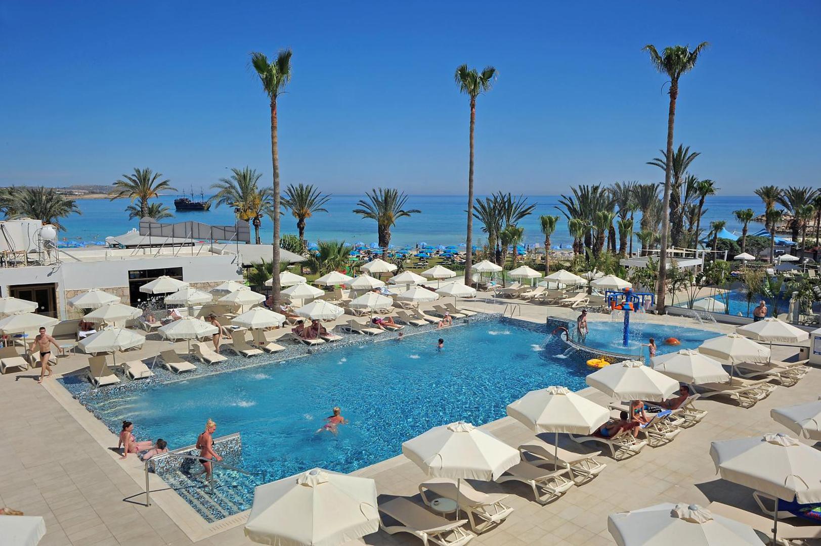 Nelia Beach Hotel galvenais baseins ar skatu uz Vidusjūru Aijanapā — saulessargi, zviļņi un palmas piekrastes joslā