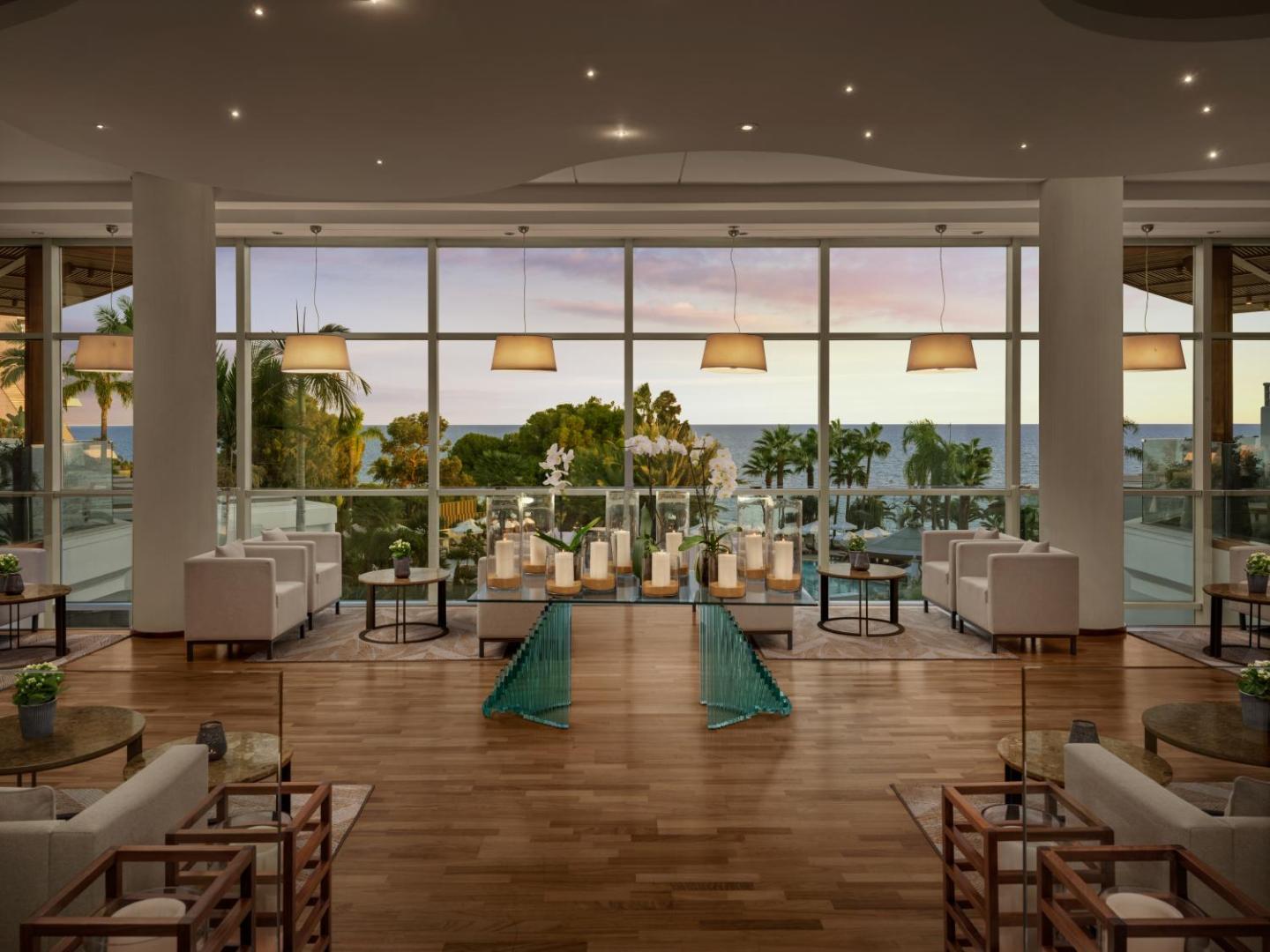 Mediterranean Beach Hotel lobby bārs ar lieliem panorāmas logiem, skatu uz Vidusjūru saulrietā un mājīgām atpūtas mēbelēm.