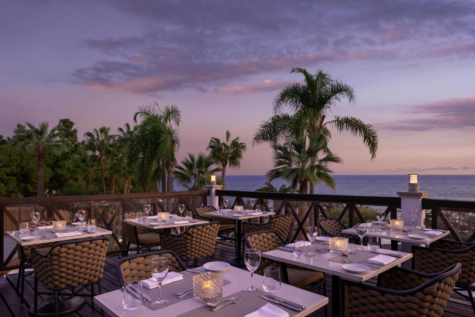 Mediterranean Beach Hotel āra terases restorāns saulrietā ar skatu uz Vidusjūru, palmām un eleganti klātiem galdiem
