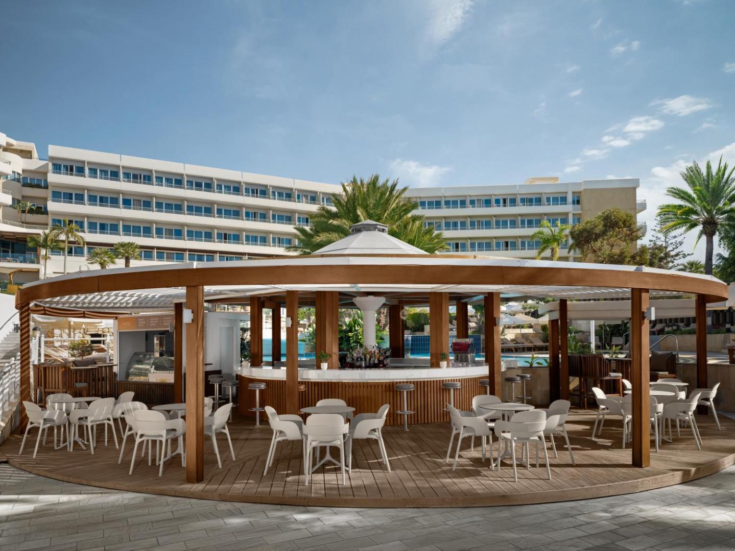 Mediterranean Beach Hotel āra bārs ar koka apdari un baltiem krēsliem pie baseina, fonā redzama viesnīcas galvenā ēka