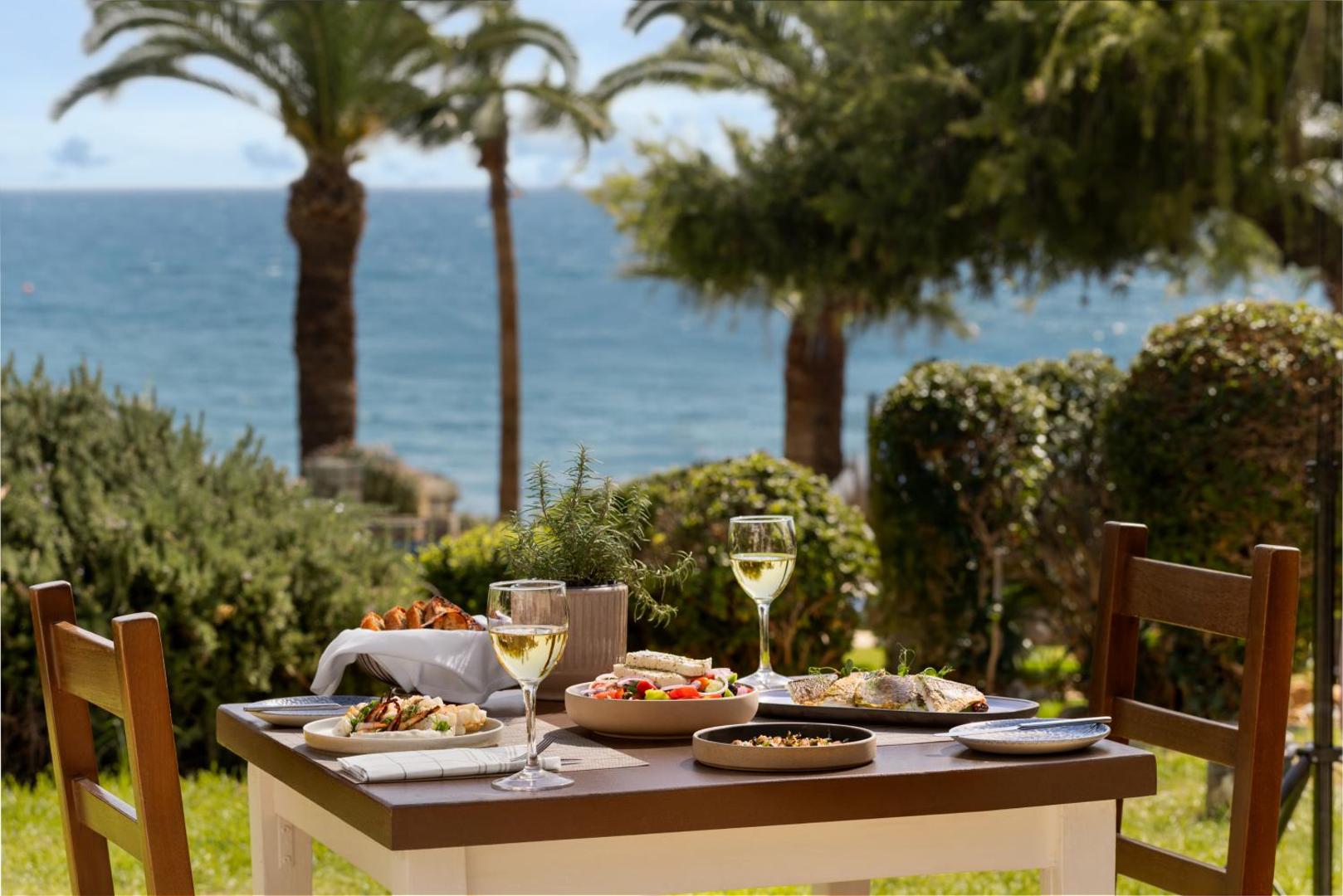 Klāts galds ar Vidusjūras virtuves ēdieniem un vīna glāzēm Mediterranean Beach Hotel dārzā pie jūras un palmām