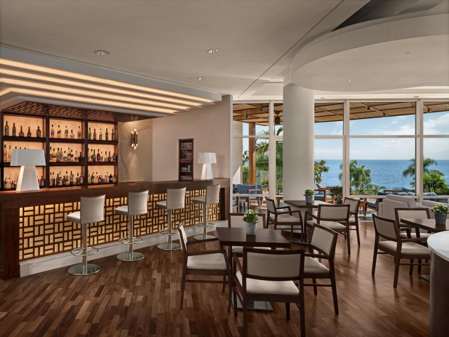 Mediterranean Beach Hotel lobija bārs ar koka apdari un skatu uz jūru Limasolā, Kiprā — eleganta atpūtas zona ar galdiņiem