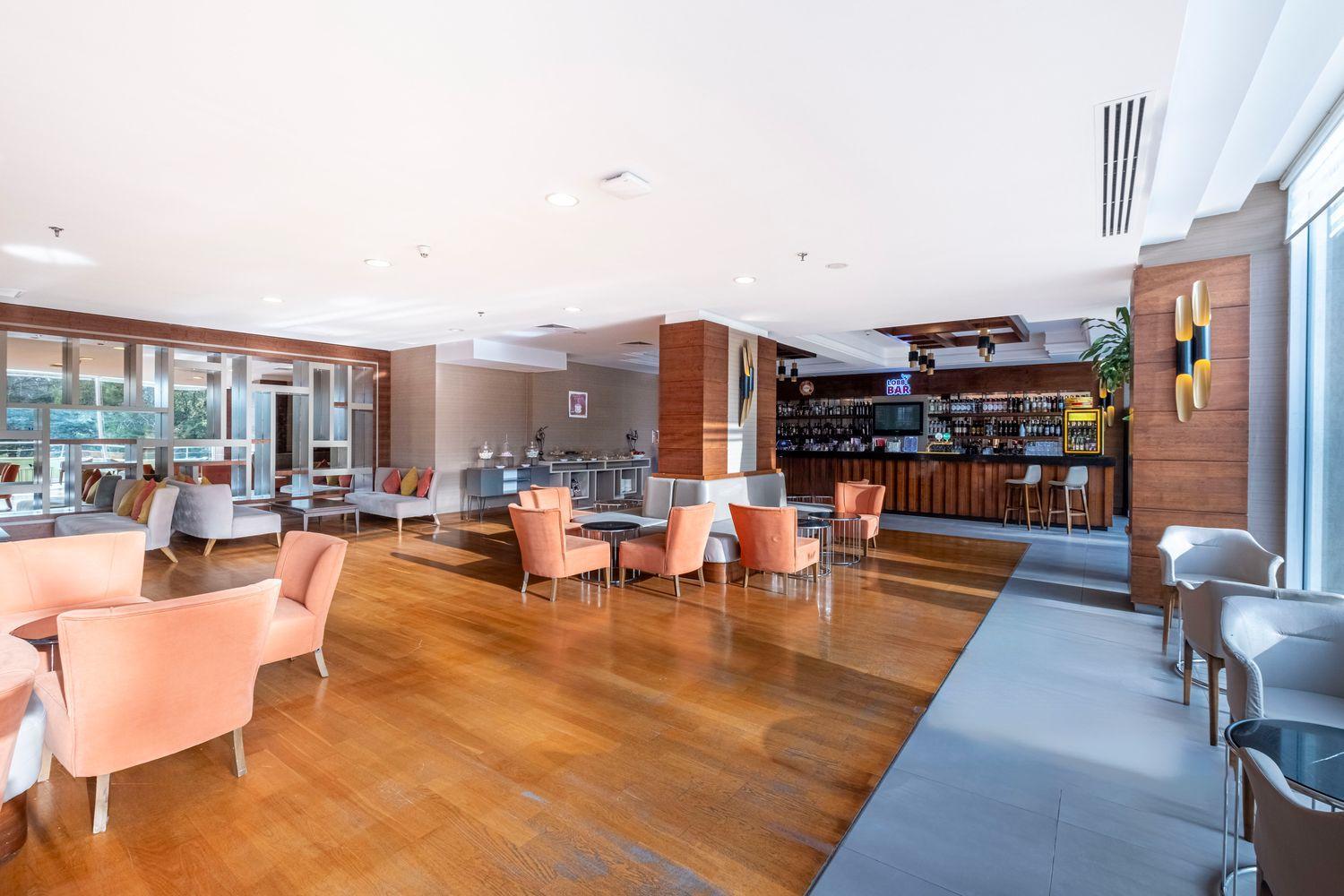 Mūsdienīgs viesnīcas vestibila bārs ar koka grīdām un ērtiem persiku krāsas atpūtas krēsliem.