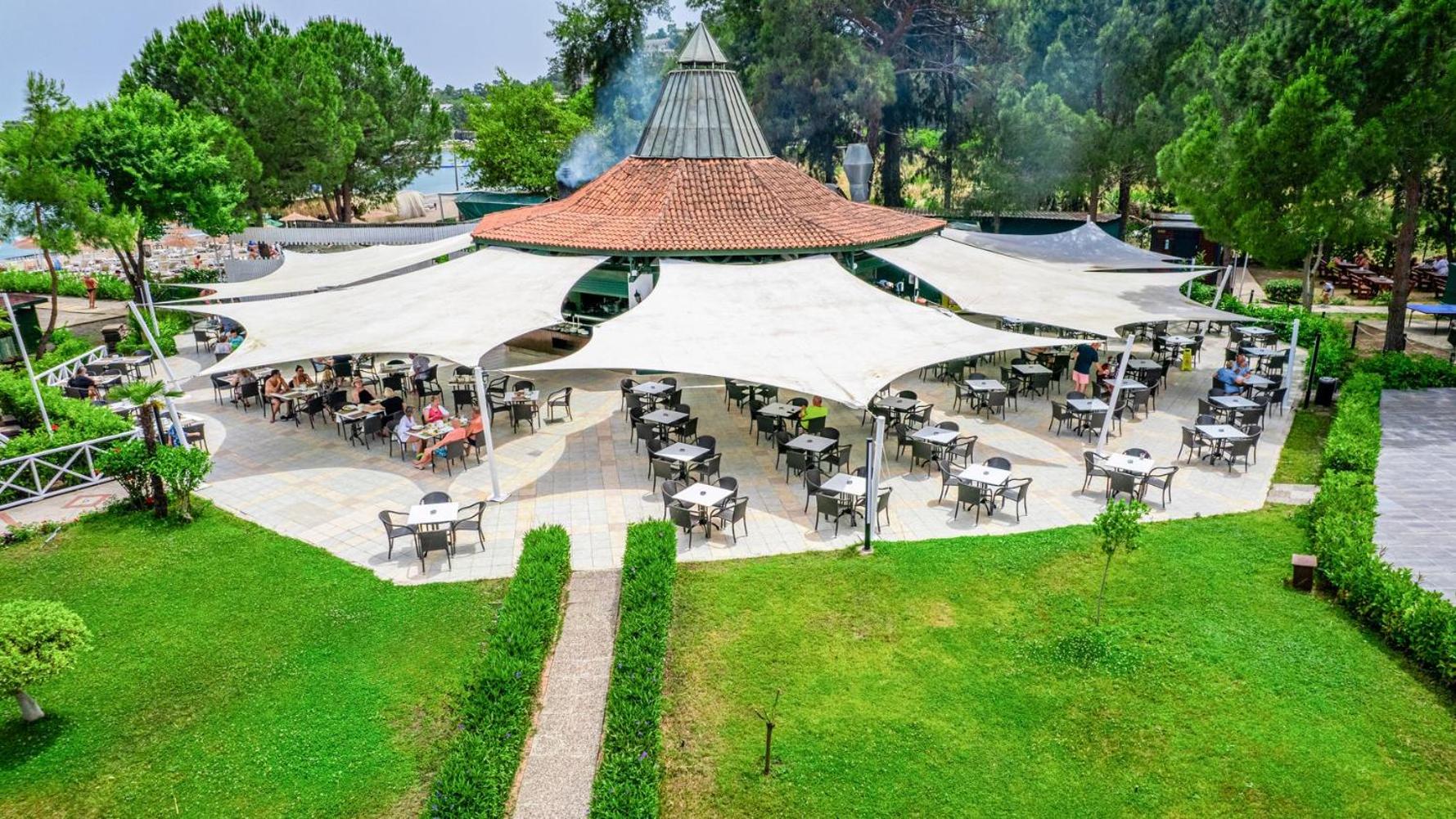 Āra bārs ar lielu jumta konstrukciju un galdiņiem, ko ieskauj kopti zālieni.