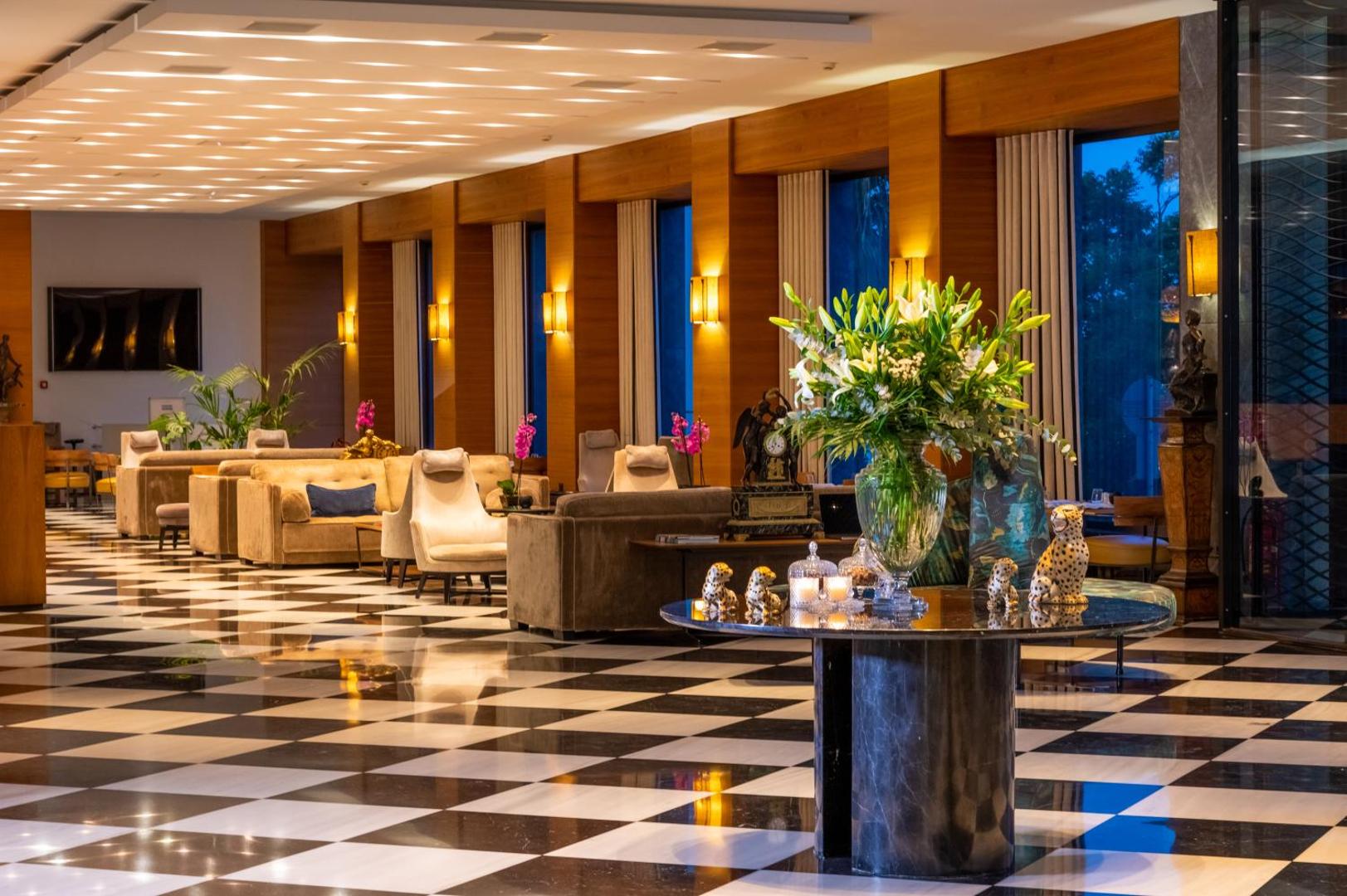 Plašs viesnīcas vestibils ar melnbaltu rūtainu grīdu, dīvāniem un ziedu kompozīciju.