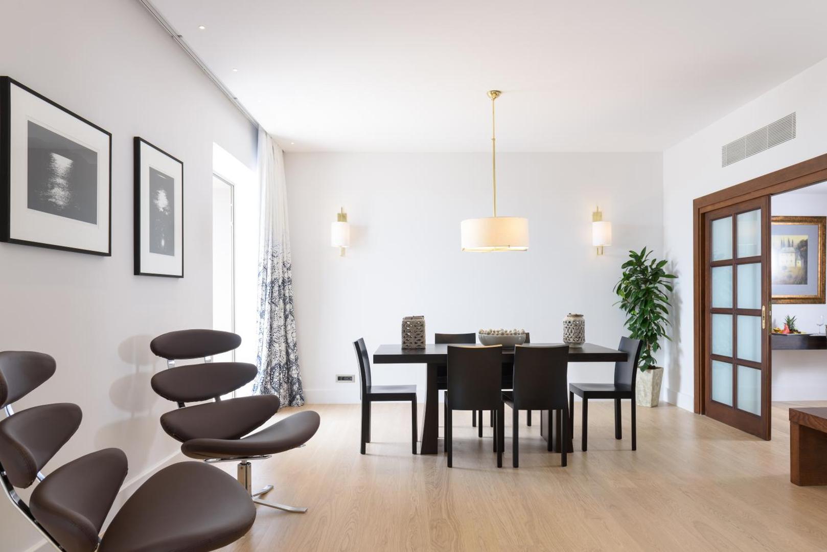 Gaiša viesnīcas numura ēdamistabas zona ar tumšu koka galdu un moderniem krēsliem.