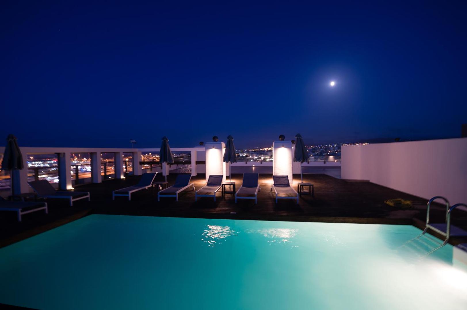 Izgaismots jumta baseins naktī zem pilnmēness ar pilsētas gaismām fonā.