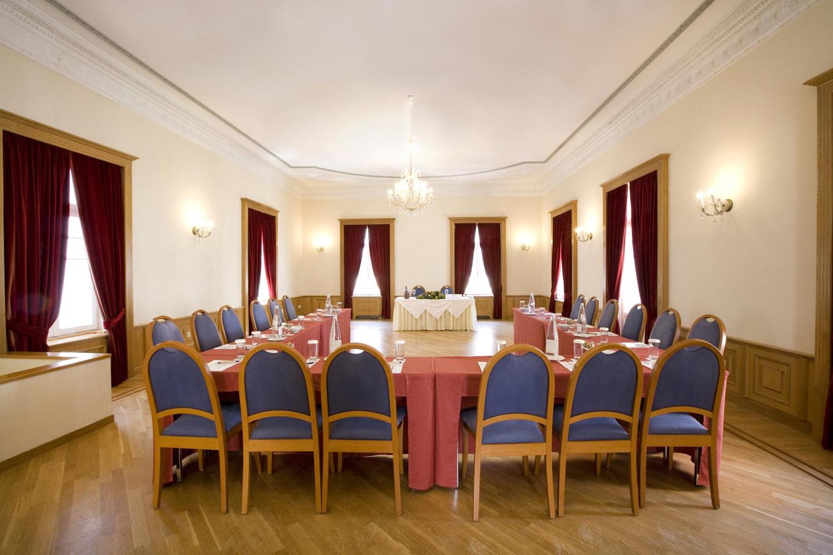 Klasiska stila sanāksmju telpa ar koka grīdu, kristāla lustru un sarkaniem aizkariem.