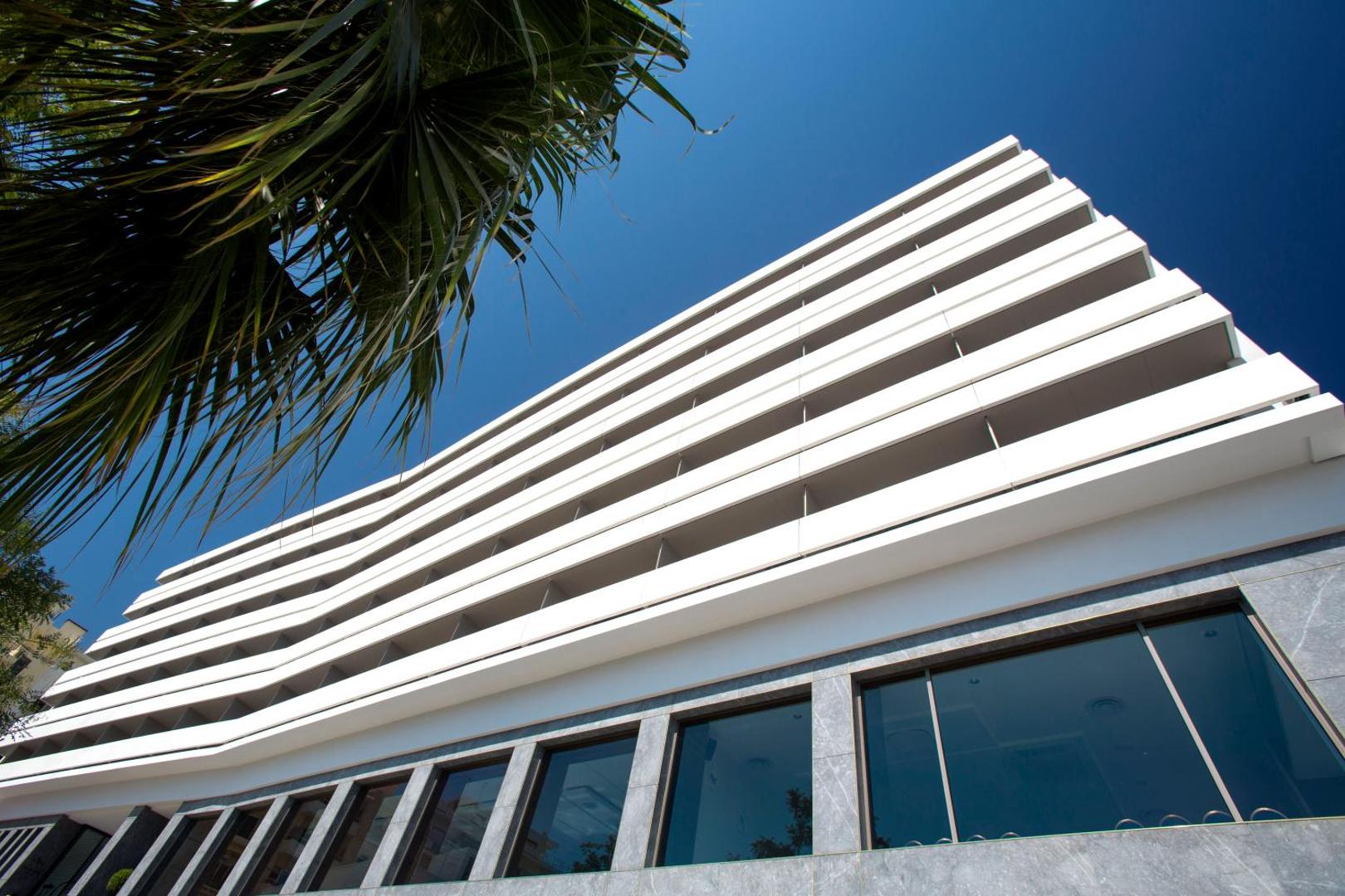 Skats no apakšas uz viesnīcas balto ēku un palmu zariem saulainā dienā.
