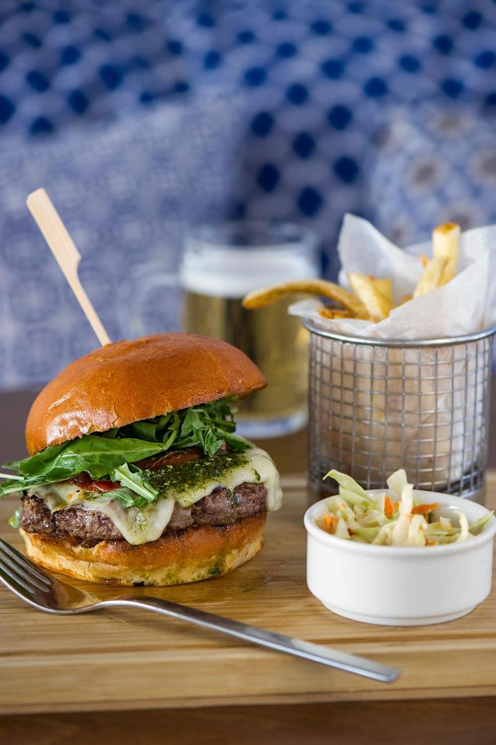 Grecian Sands restorāna piedāvājums — sulīgs burgers ar pesto, frī kartupeļi un salāti pasniegti uz koka dēlīša