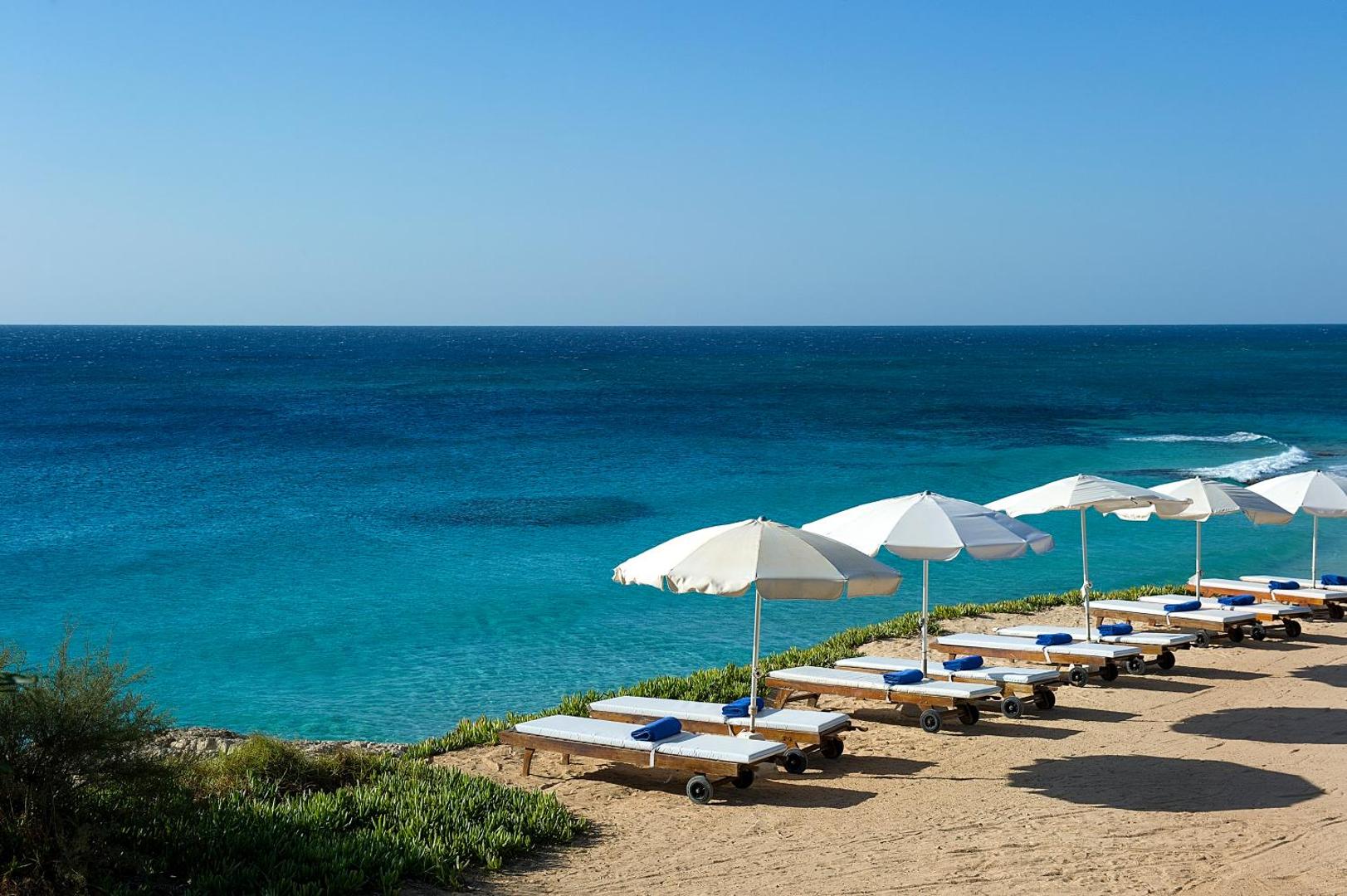 Grecian Sands privātā pludmales zona ar sauļošanās krēsliem un saulessargiem uz klints malas pie tirkīzzilas jūras