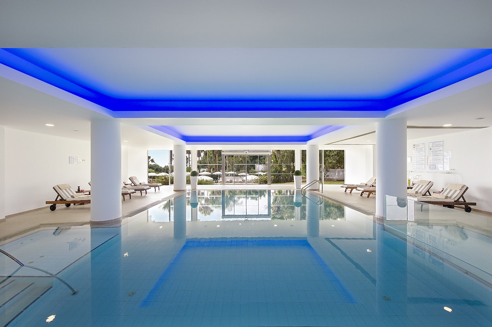 Grecian Sands spa centra iekštelpu baseins ar zilu neona apgaismojumu — atpūtas krēsli un skats caur lielajiem logiem uz dārzu