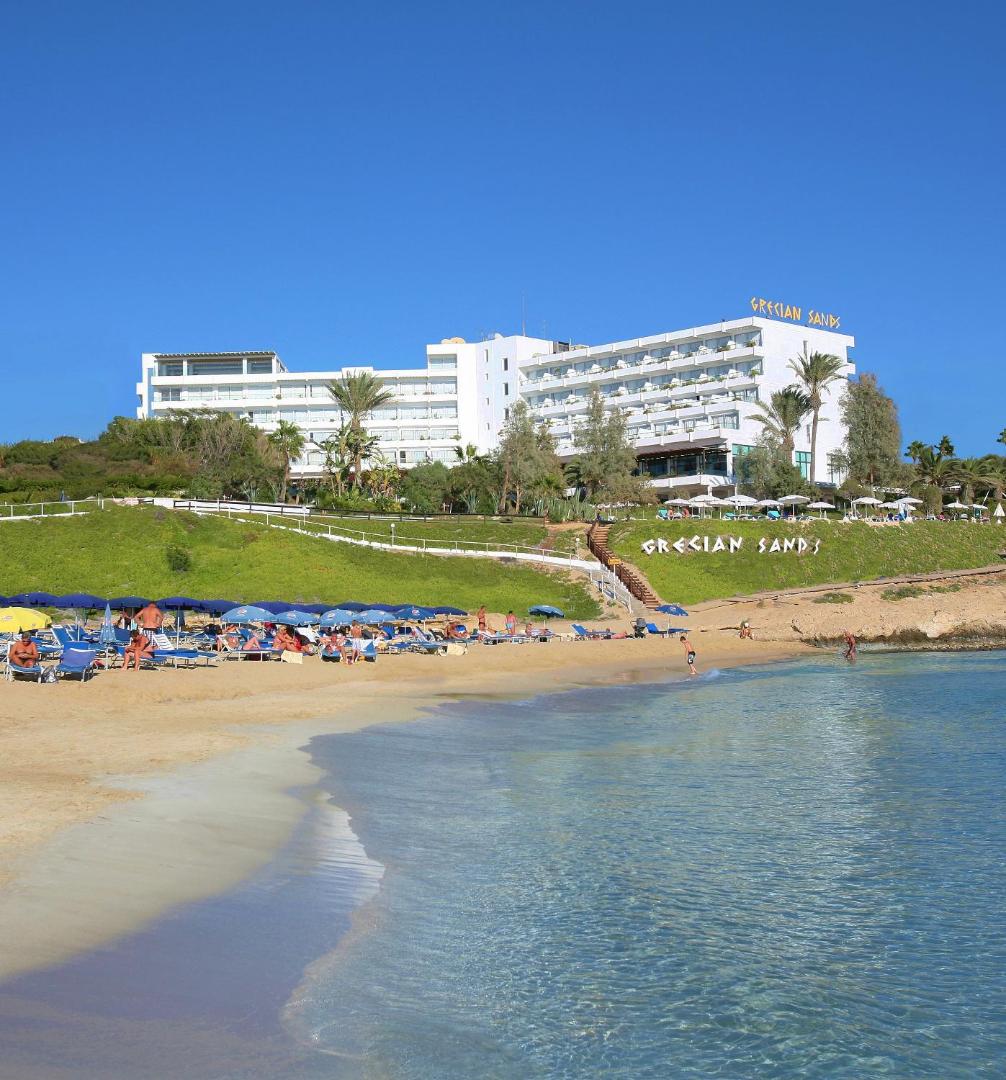 Grecian Sands viesnīcas ēka skatā no pludmales ar saulessargiem un dzidru jūras ūdeni Ajanapas piekrastē