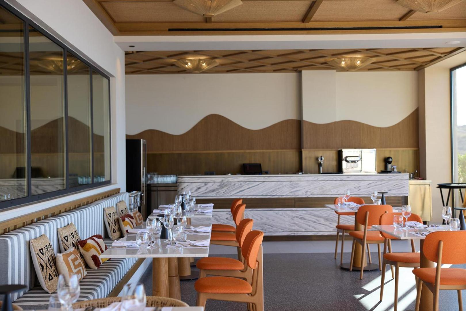 Mūsdienīgs restorāna interjers ar marmora bāru, oranžiem krēsliem un gaišām mēbelēm.