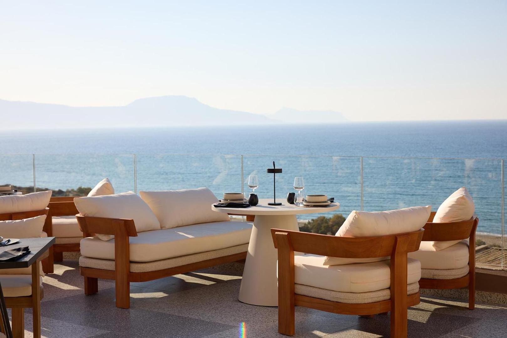 Āra terase ar koka atpūtas krēsliem un galdiņu piekrastē ar skatu uz jūru.