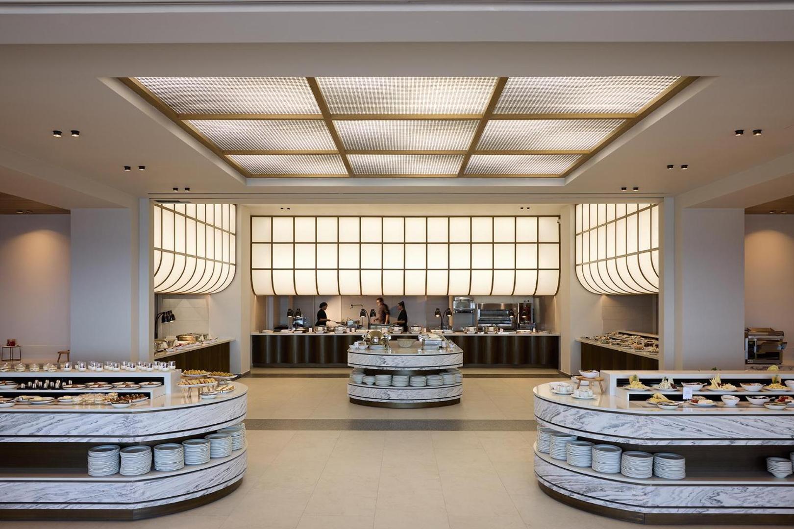 Mūsdienīga viesnīcas bufetes zona ar marmora letēm un siltu, ģeometrisku griestu apgaismojumu.