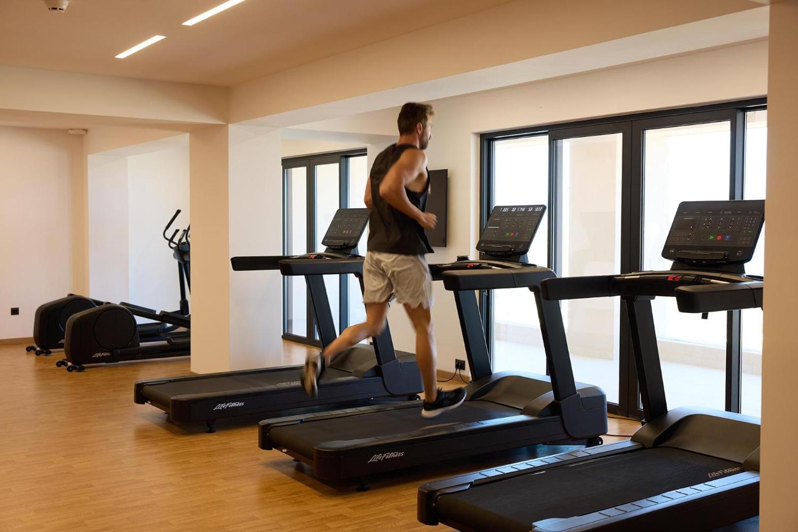 Vīrietis sporta tērpā skrien uz skrejceliņa gaišā un modernā viesnīcas sporta zālē.